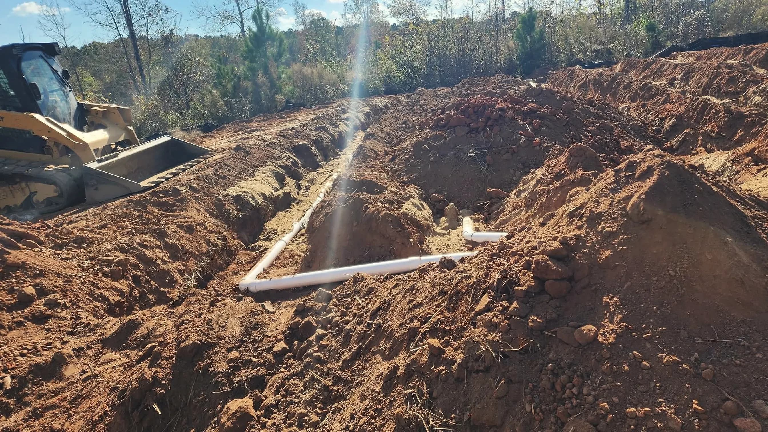 From our job files: drain field trenches with white PVC pipe cut into red clay beside a skid steer on a wooded lot; illustrating septic repair for Covington, Georgia