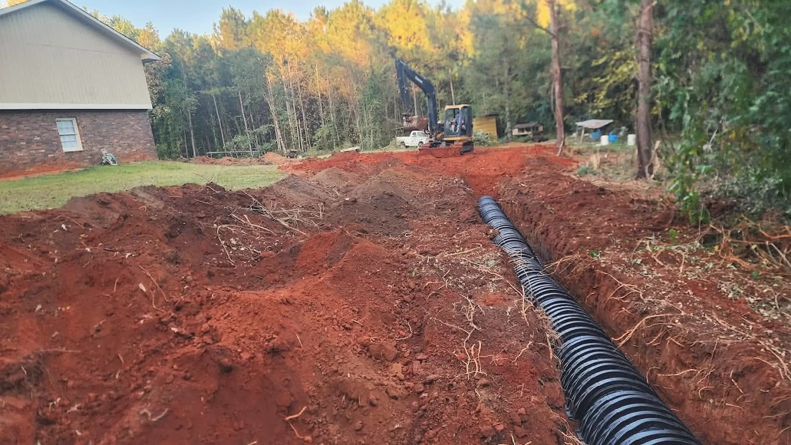 From our job files: a long trench with black corrugated pipe beside a brick house, an excavator working behind it; illustrating sewer line repair for Lithonia, Georgia