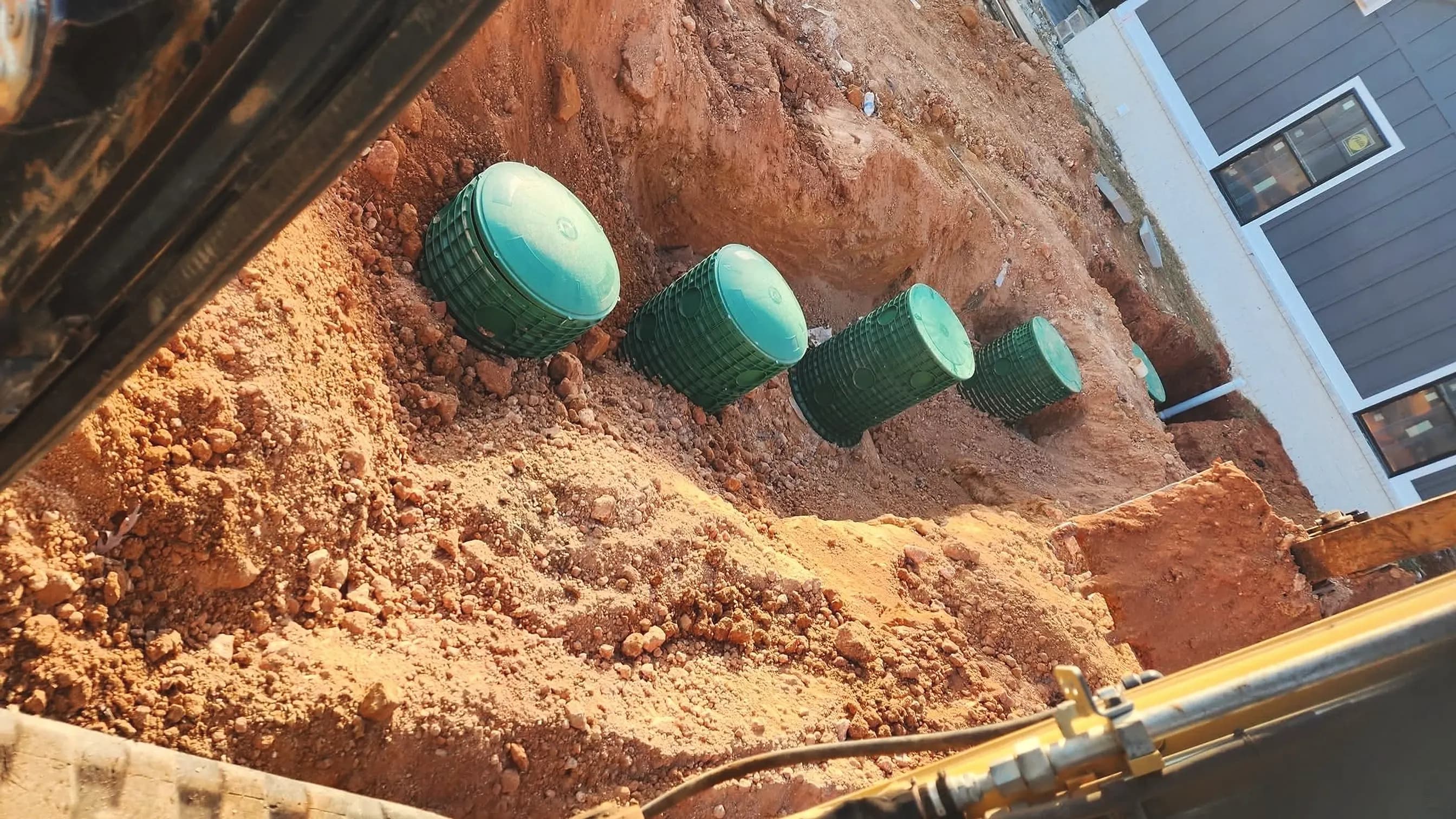 From our job files: a row of green septic riser lids in a fresh excavation beside a new home, seen from the excavator cab; illustrating septic repair for Rutledge, Georgia